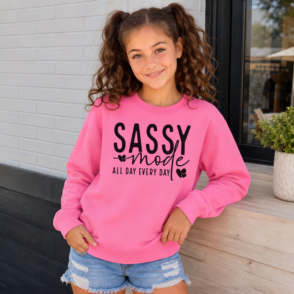 Youth girl wearing a bright pink Sassy Mode sweatshirt with bold black lettering outside a storefront.