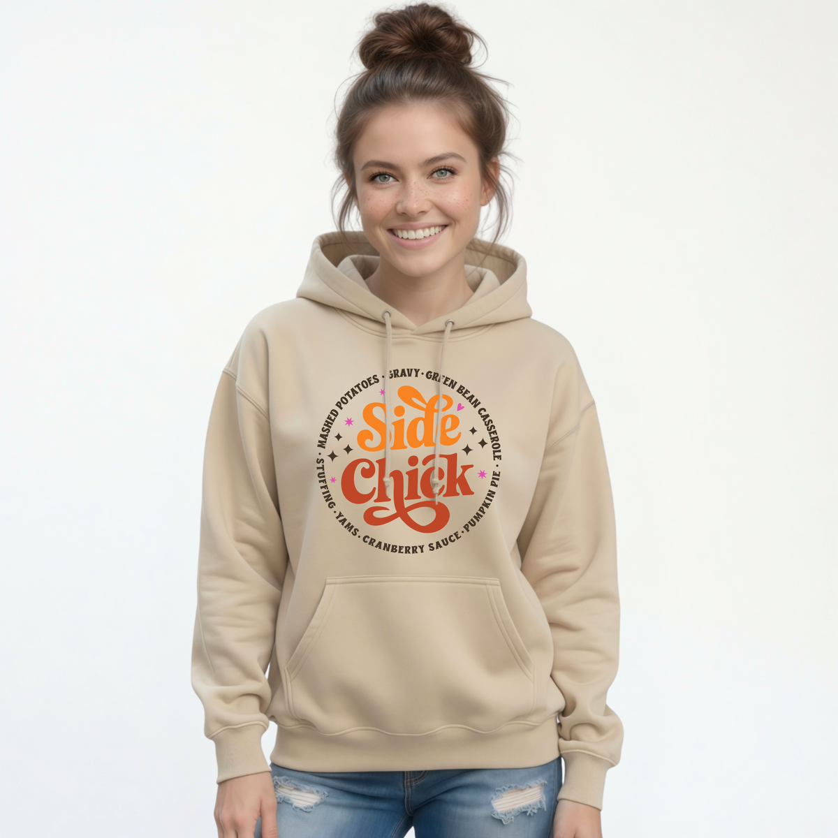 A smiling woman with her hair in a bun wears a Sand pullover hoodie featuring the retro “Side Chick” Thanksgiving graphic with dark side-dish lettering, paired with blue jeans on a white background.