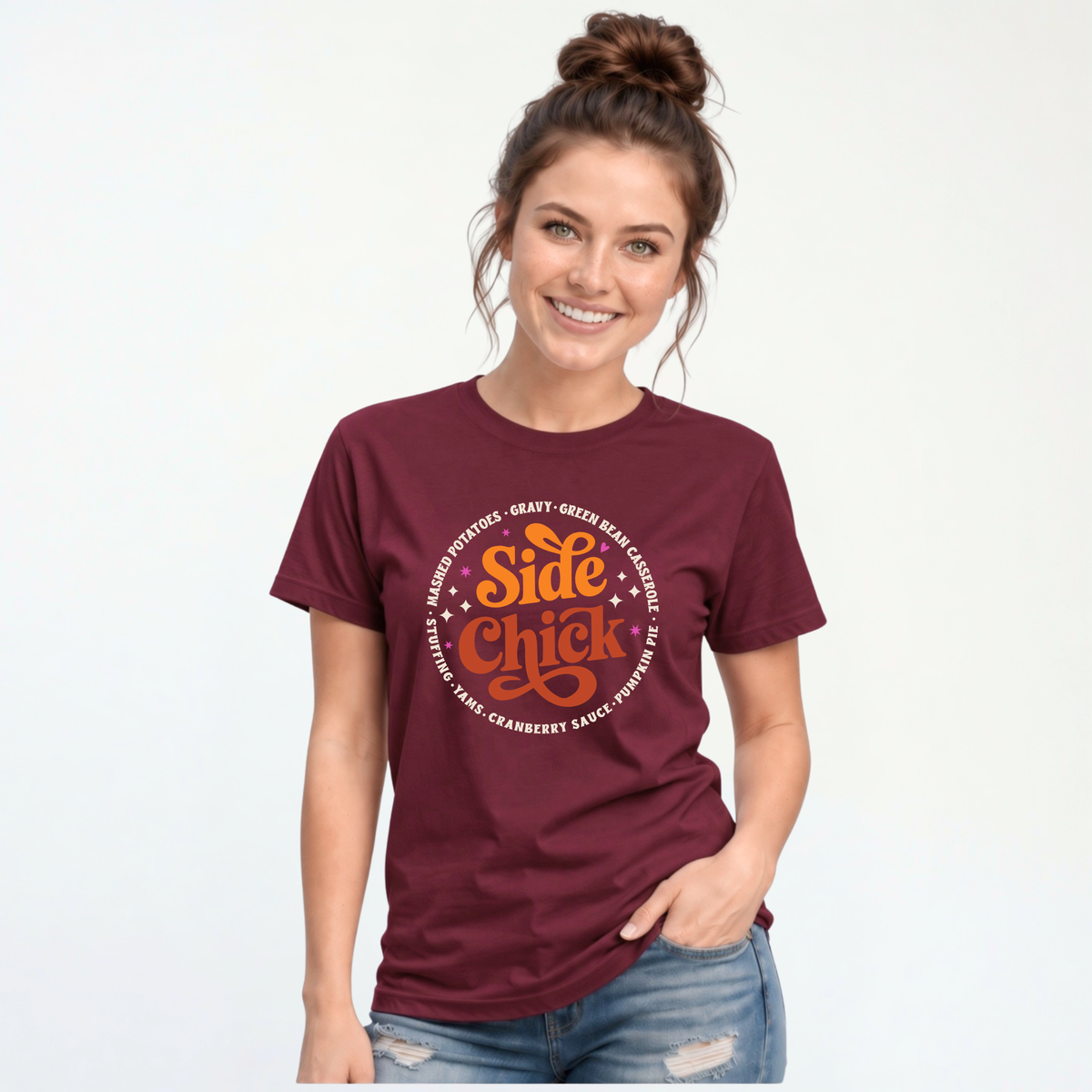 A woman wears a Maroon short-sleeve T-shirt featuring the retro “Side Chick” Thanksgiving design with bright side-dish lettering, photographed against a white studio background.
