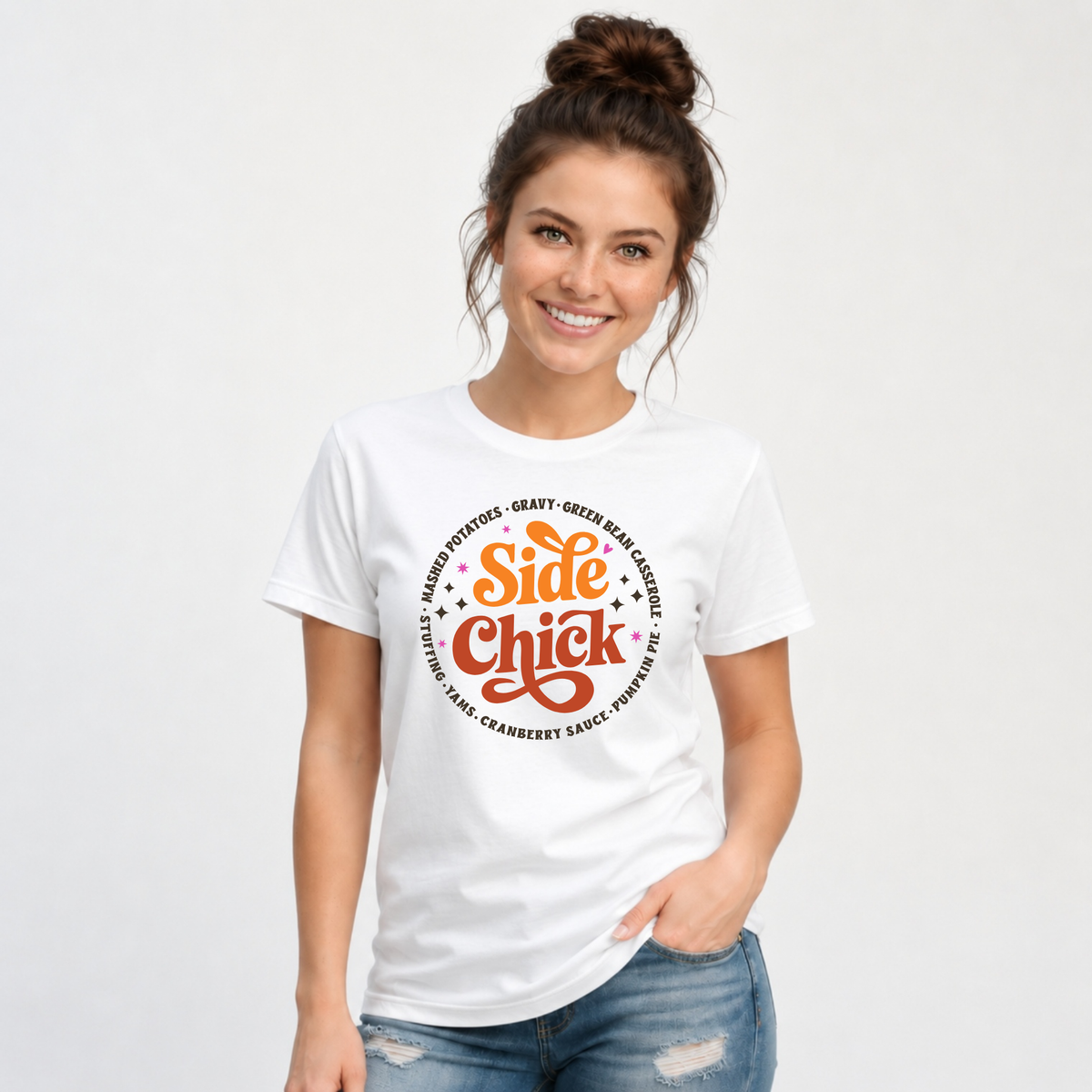 A woman models a White T-shirt with the orange-and-rust “Side Chick” Thanksgiving graphic and dark circular side-dish lettering, paired with jeans in a bright studio setting.