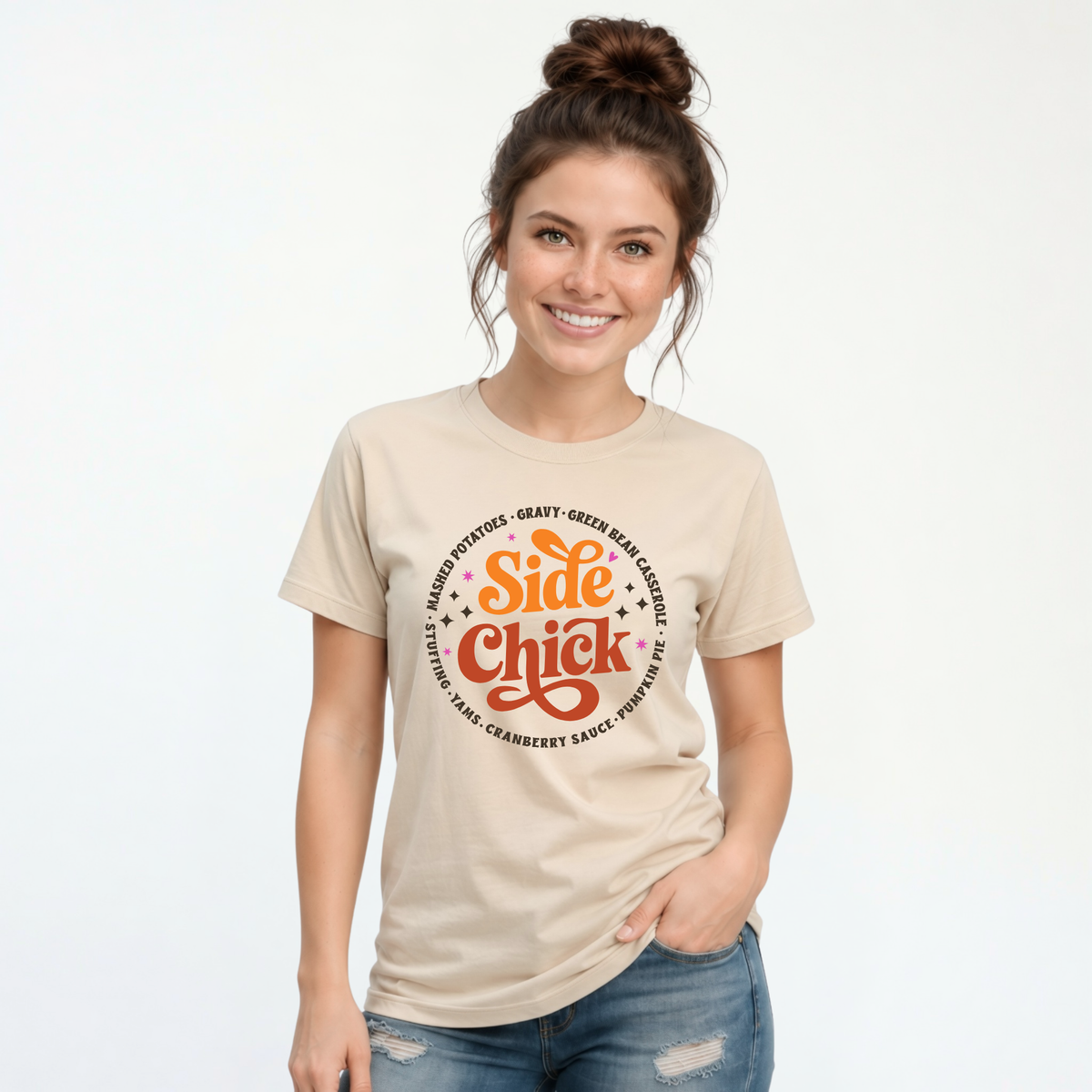 A woman models a Sand T-shirt with a retro “Side Chick” Thanksgiving graphic, featuring orange and rust lettering surrounded by side-dish names, against a clean white background.