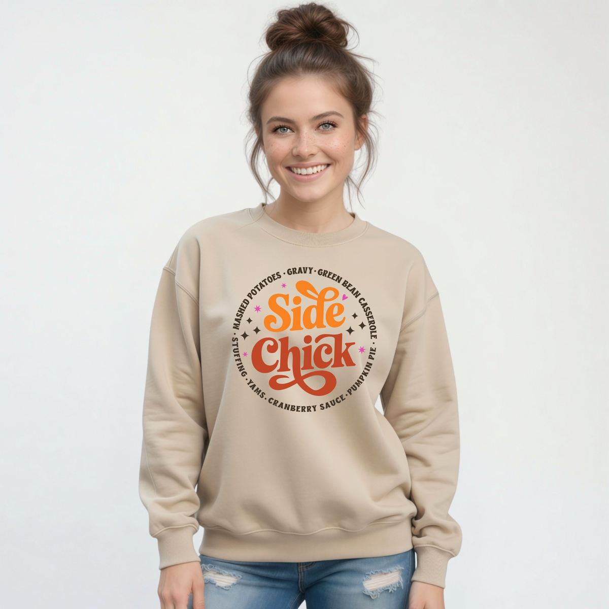 A woman wears a Sand crewneck sweatshirt with the retro “Side Chick” Thanksgiving design, surrounded by dark lettering naming classic holiday side dishes, on a light studio background.