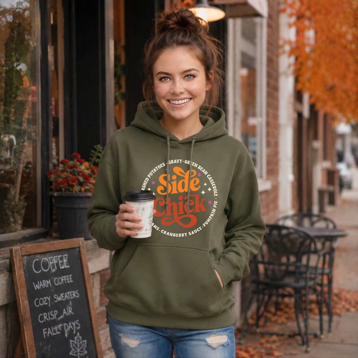 A woman wears a Military Green “Side Chick” Thanksgiving hoodie on a sidewalk outside a coffee shop, giving the funny fall design a casual lifestyle look.