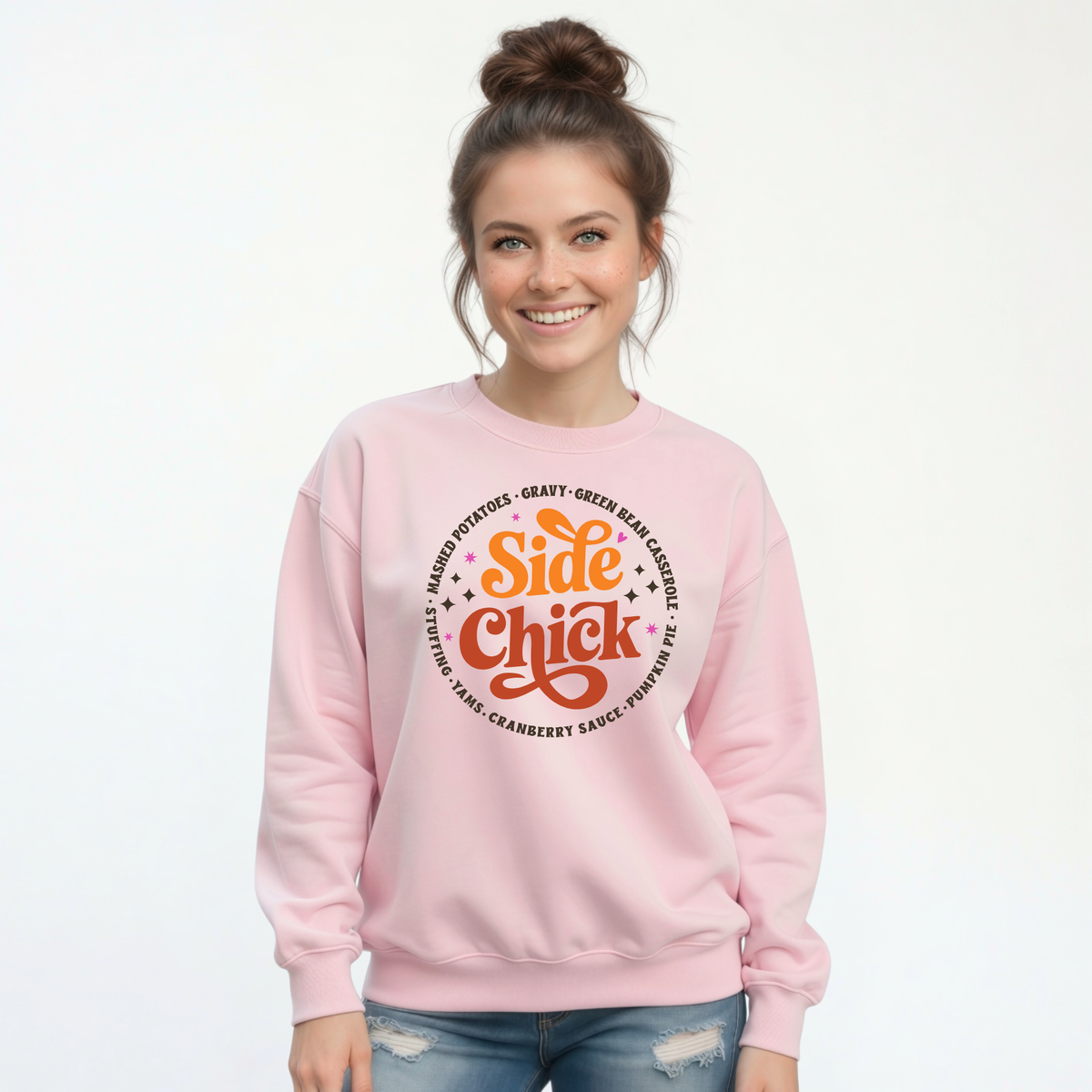 A smiling woman with her hair in a bun wears a Light Pink crewneck sweatshirt featuring the retro “Side Chick” Thanksgiving graphic, dark side-dish lettering, and small star accents against a white background.