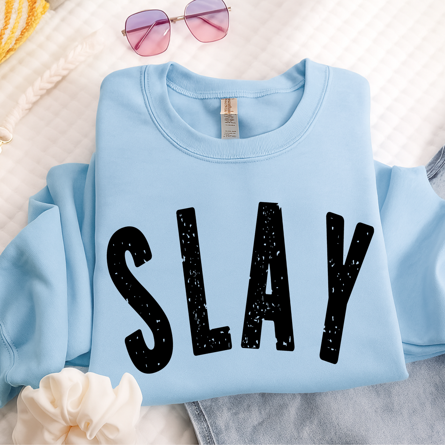Light blue sweatshirt with 'SLAY' text on a white surface with sunglasses and flowers.