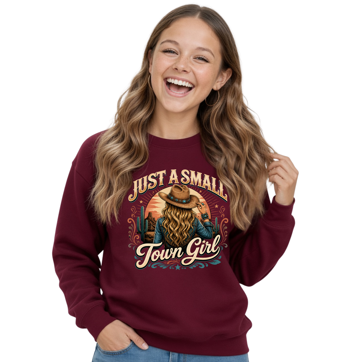 A smiling young girl with long wavy brown hair wears a maroon crewneck sweatshirt featuring a western-themed graphic with the text 'JUST A SMALL Town Girl' and an illustration of a woman with a large hat and long hair. She is wearing blue jeans against a plain white background.