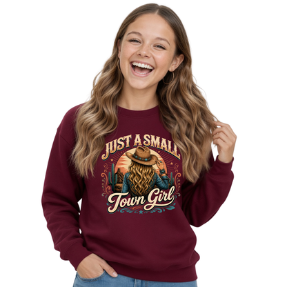 A smiling young girl with long wavy brown hair wears a maroon crewneck sweatshirt featuring a western-themed graphic with the text 'JUST A SMALL Town Girl' and an illustration of a woman with a large hat and long hair. She is wearing blue jeans against a plain white background.