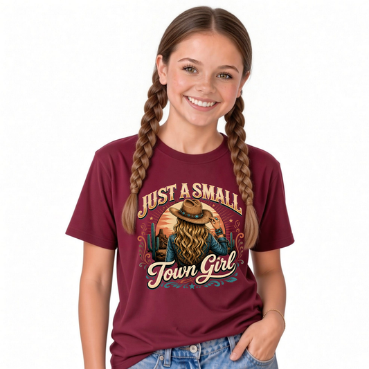 A young girl with long braids is wearing a maroon graphic t-shirt with a western-themed design that reads 'JUST A SMALL Town Girl' and shows a cowboy with a hat and  long blonde hair. She is also wearing blue jeans against a plain white background.