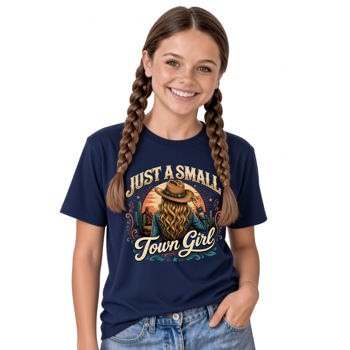 A smiling girl with long braided hair wears a navy blue graphic T-shirt featuring a cowboy illustration and the text 'JUST A SMALL Town Girl' in cream and tan lettering, paired with light blue jeans against a plain white background.