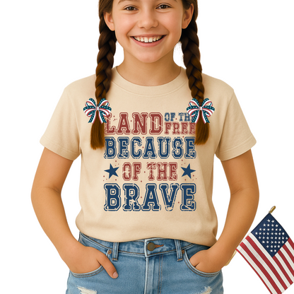 Smiling girl in sand-colored "Land of the Free Because of the Brave" shirt, styled with American flag hair bows and denim shorts