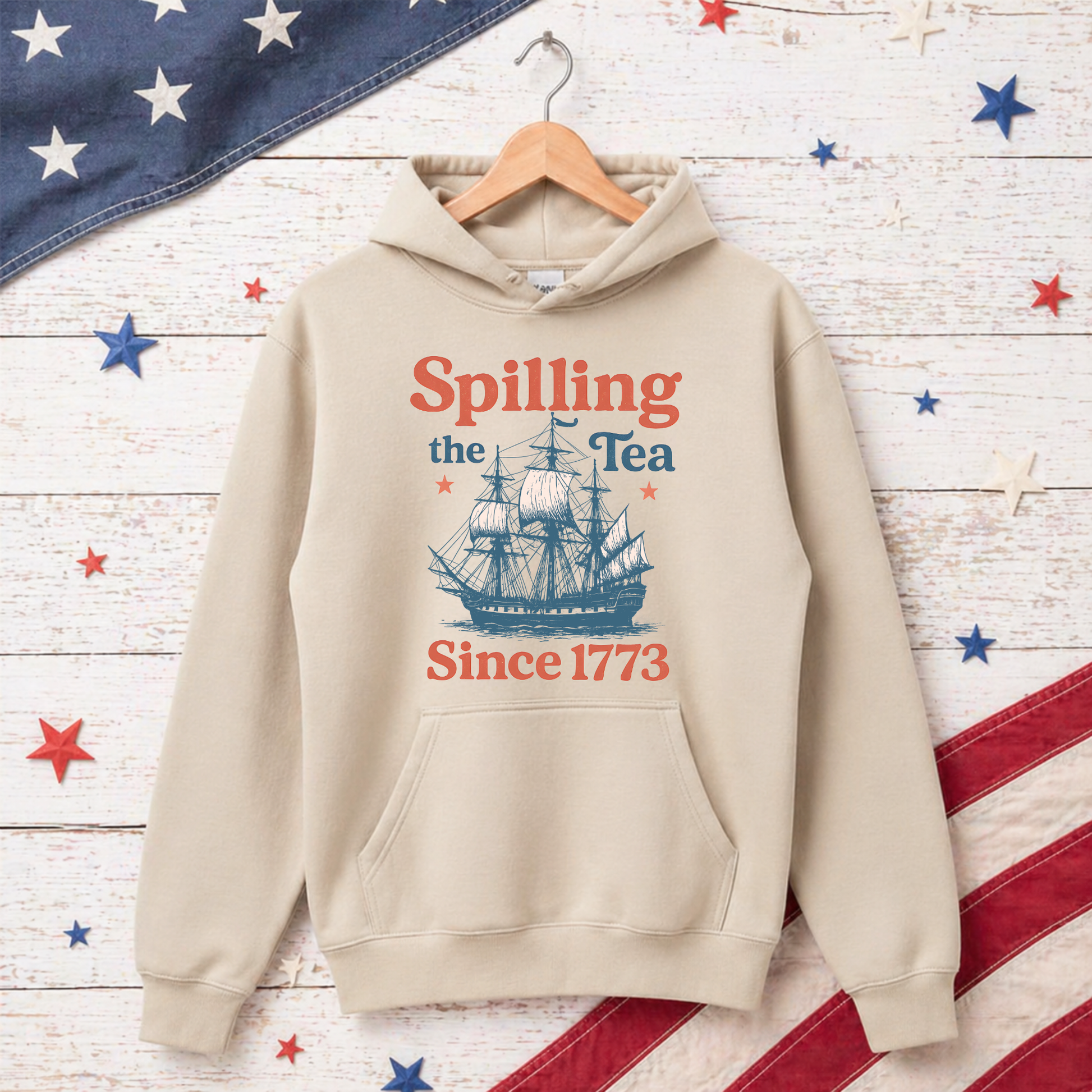 Beige hoodie with ship graphic and text 'Spilling the Tea Since 1773' on a wooden surface with American flag decorations.