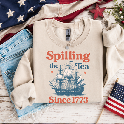 Beige sweatshirt with 'Spilling the Tea Since 1773' design on a wooden surface with American flags and denim jeans.