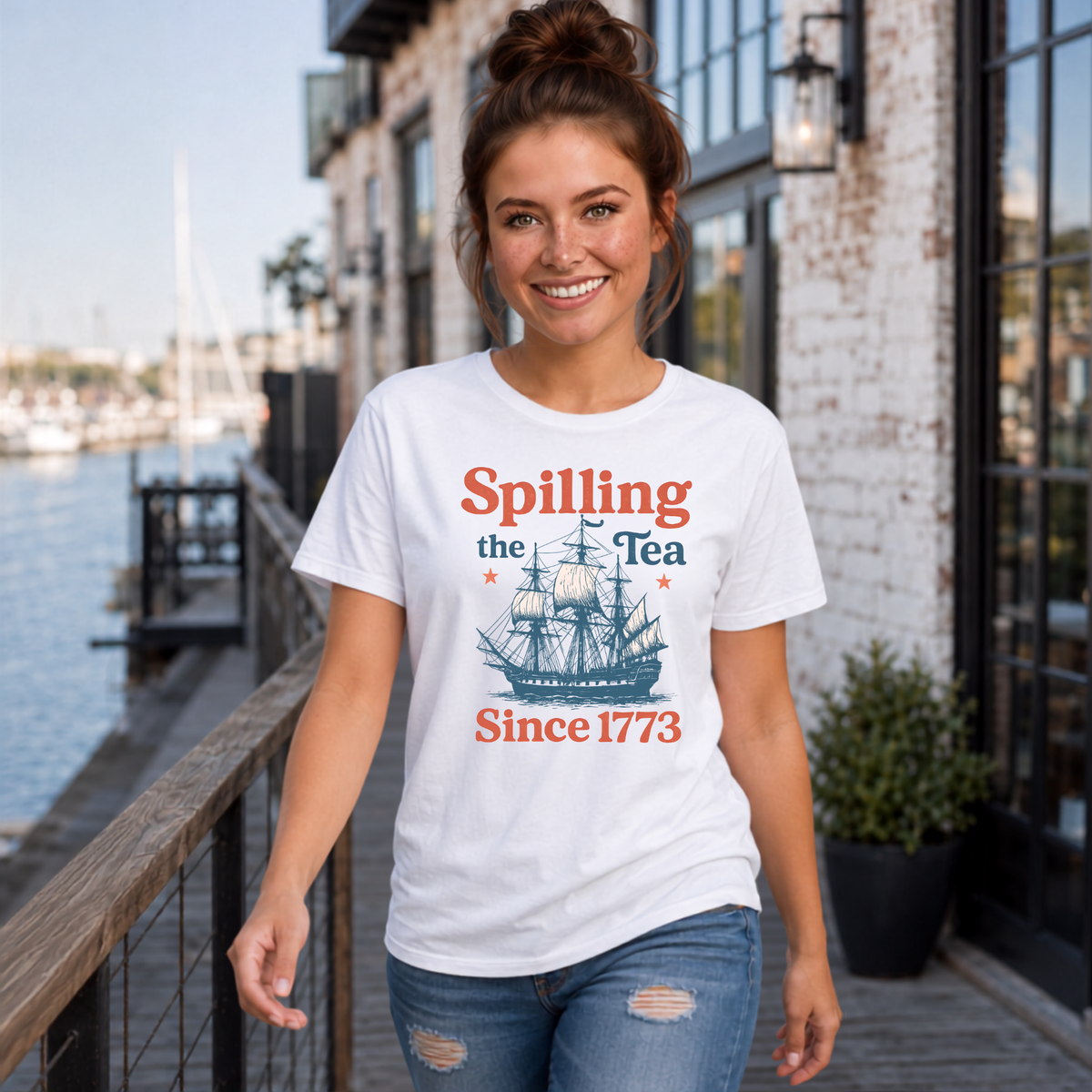 Woman wearing a white Spilling the Tea Since 1773 T-shirt with a vintage ship graphic by a marina storefront.