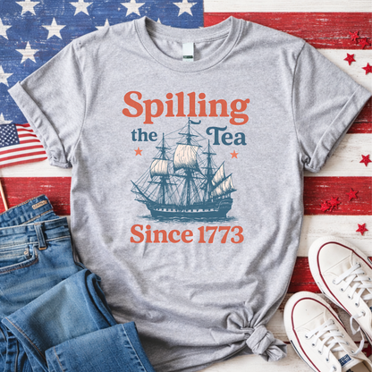 Gray t-shirt with ship graphic and text 'Spilling the Tea Since 1773' on a wooden surface with American flag and jeans.