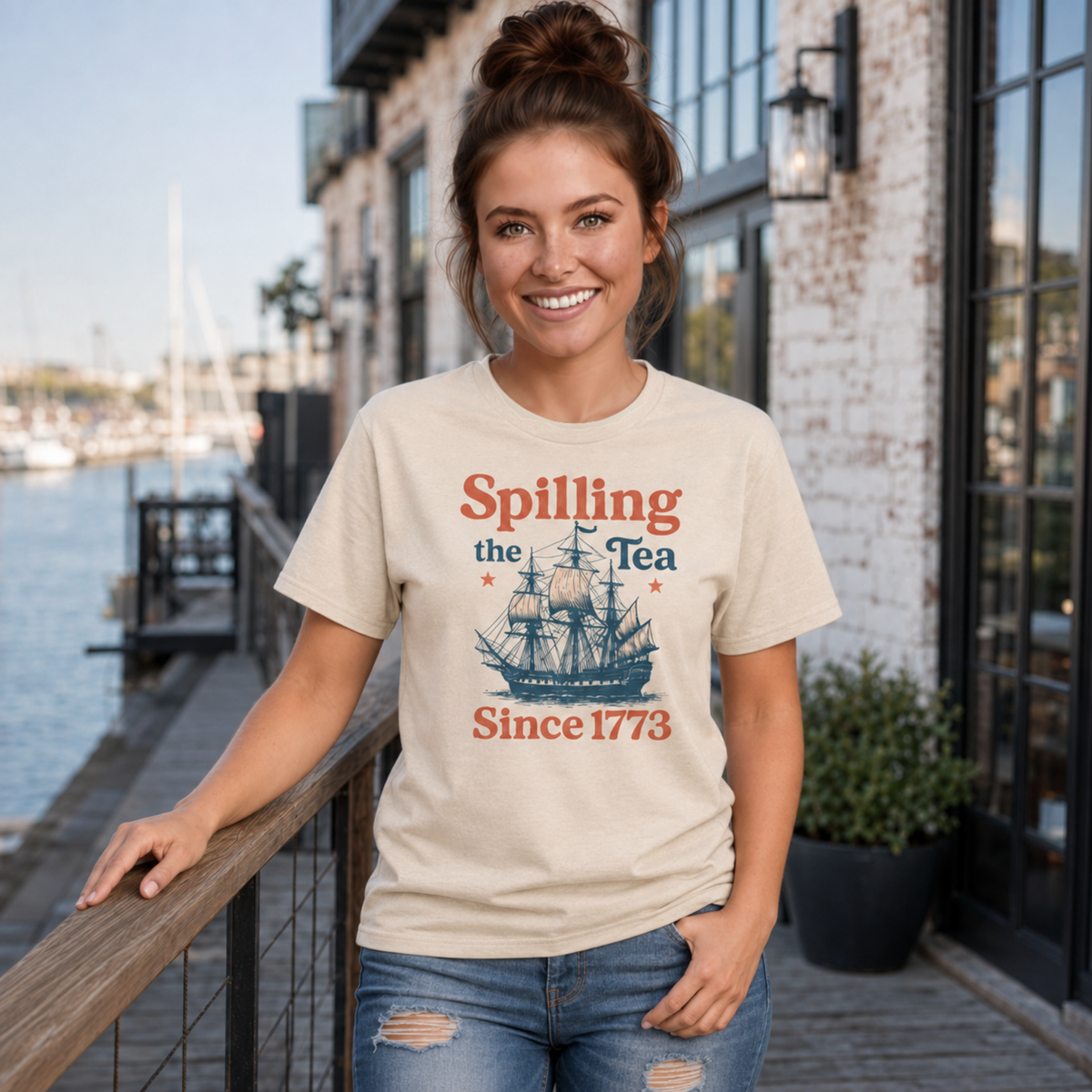 Woman wearing a sand Spilling the Tea Since 1773 T-shirt with a vintage ship graphic by a marina storefront.