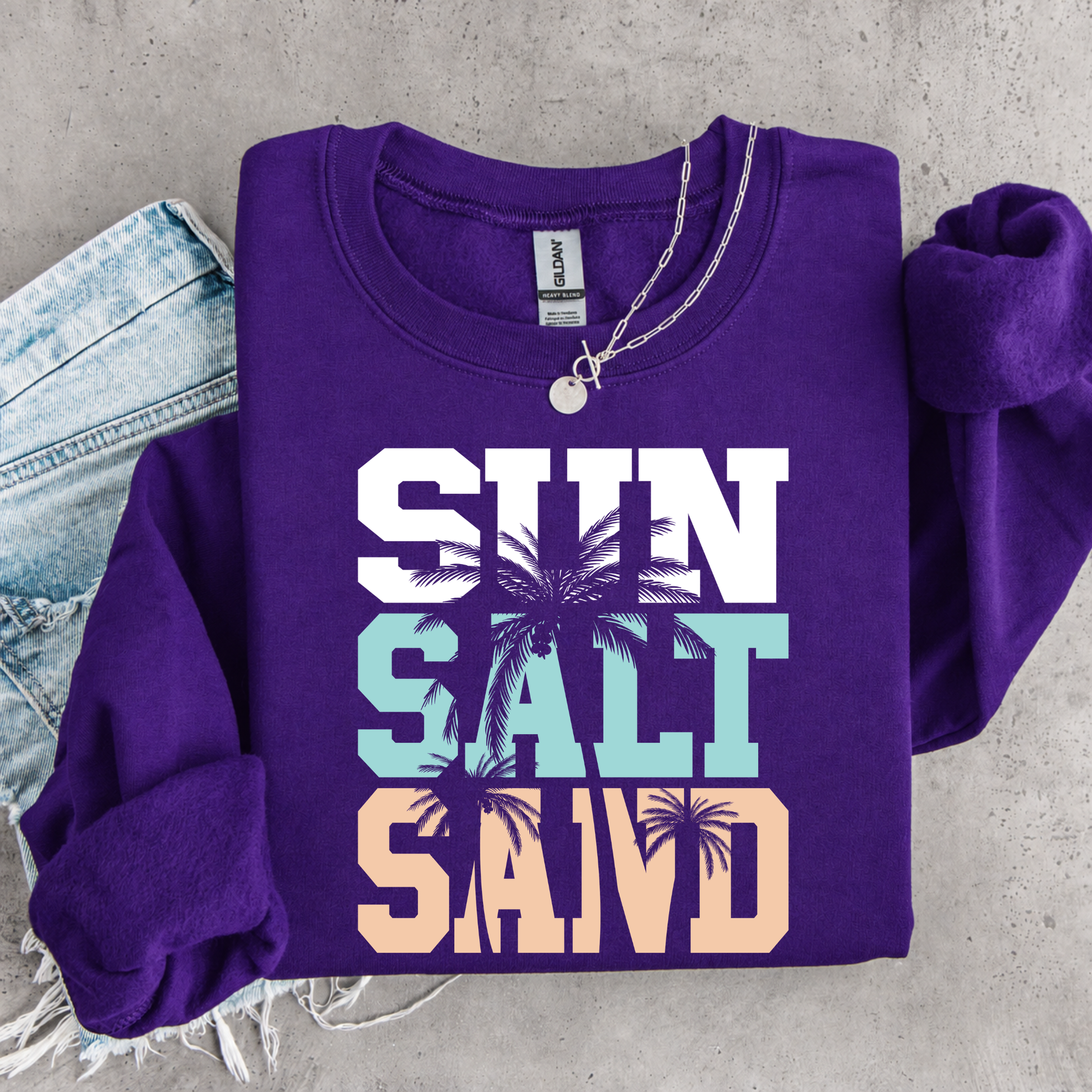 Purple sweatshirt with 'Sun Salt Sand' text and palm tree graphics on a concrete surface.