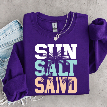 Purple sweatshirt with 'Sun Salt Sand' text and palm tree graphics on a concrete surface.