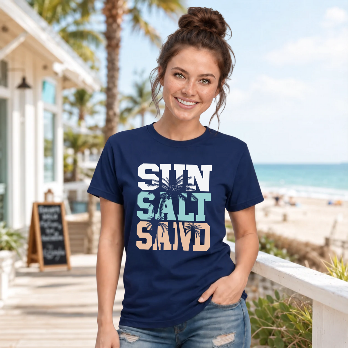 Woman wearing a navy Sun Salt Sand T-shirt on a beachside boardwalk with palm trees and the ocean behind her.