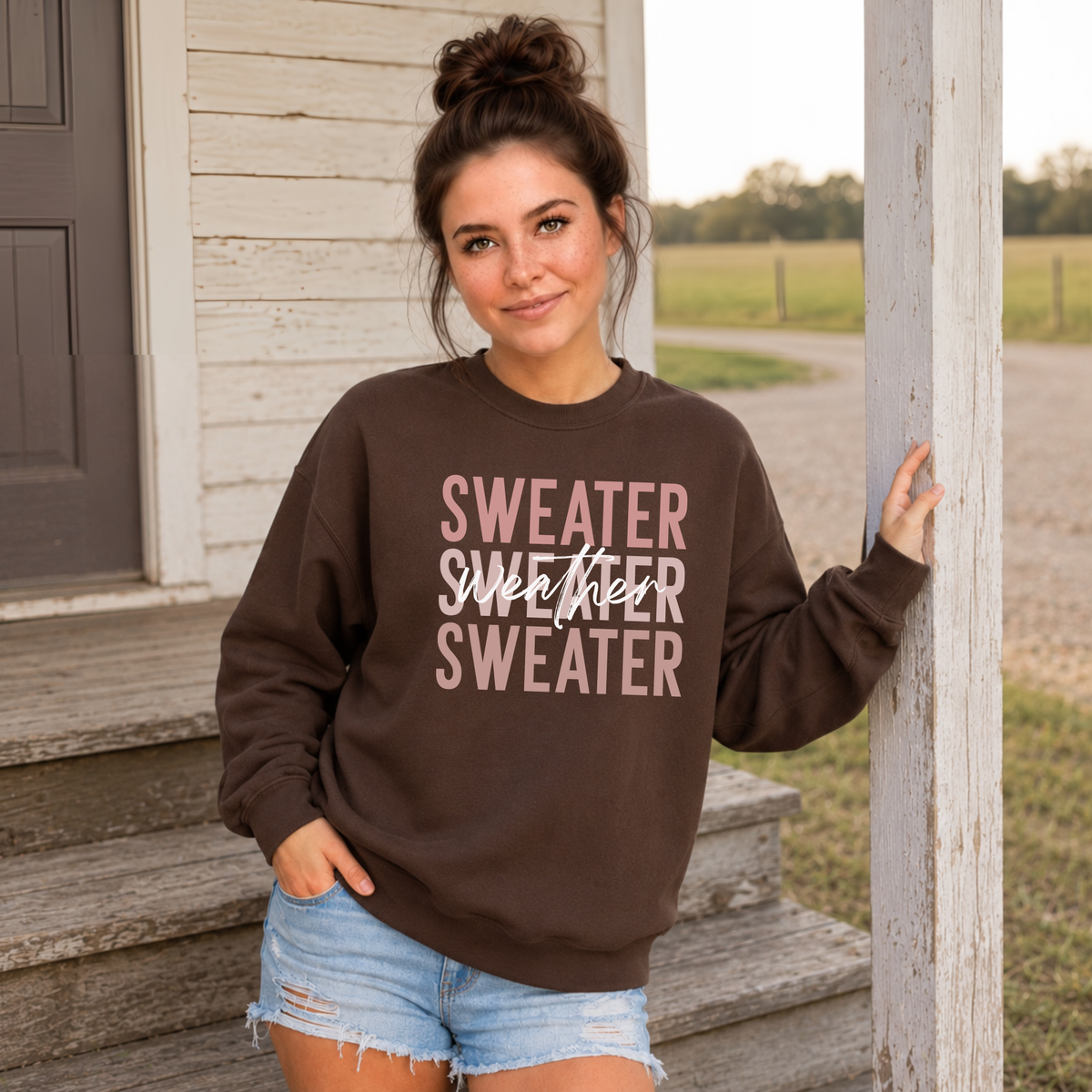 Woman standing outdoors wearing a brown “Sweater Weather Sweater” crewneck sweatshirt with denim shorts.