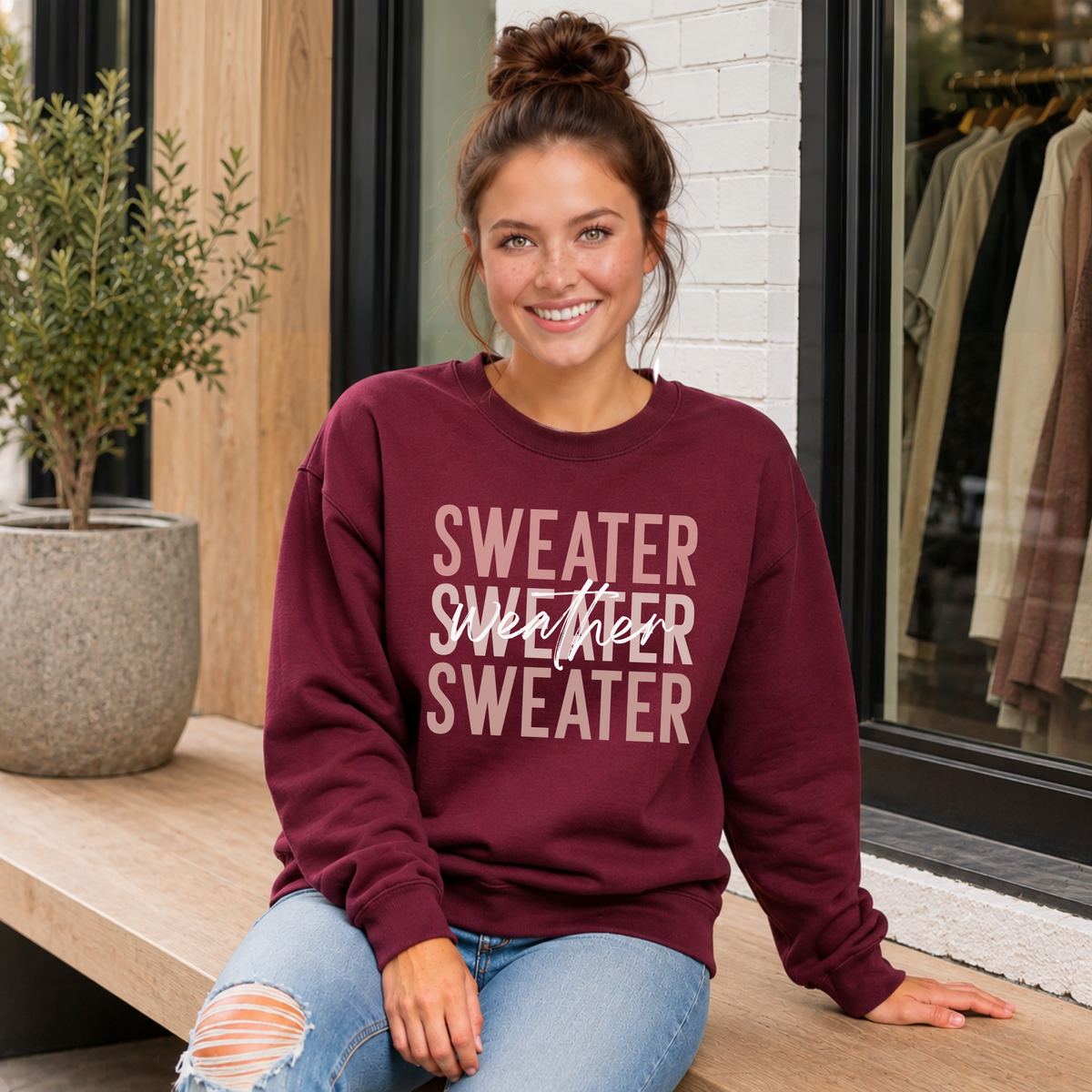 Woman sitting on a porch wearing a maroon “Sweater Weather Sweater” crewneck sweatshirt and jeans.