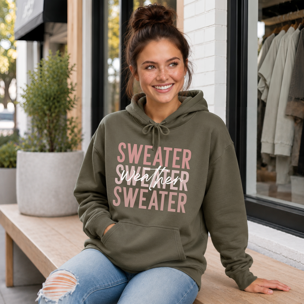 Woman sitting on a porch wearing a military green “Sweater Weather Sweater” crewneck sweatshirt and jeans.