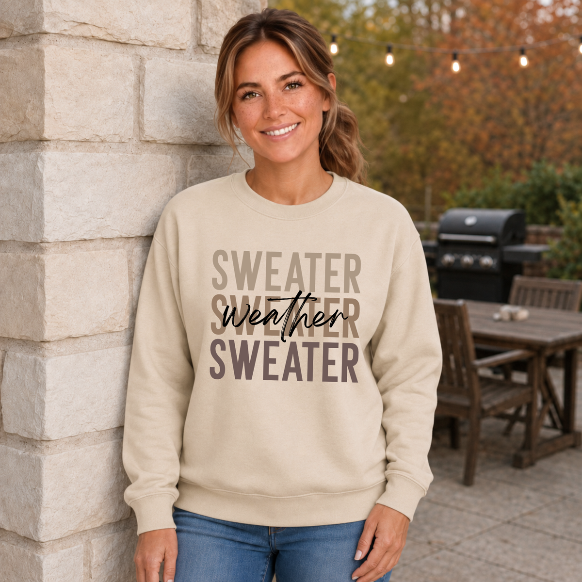 Woman outdoors wearing a sand-colored “Sweater Weather Sweater” crewneck sweatshirt with denim shorts.