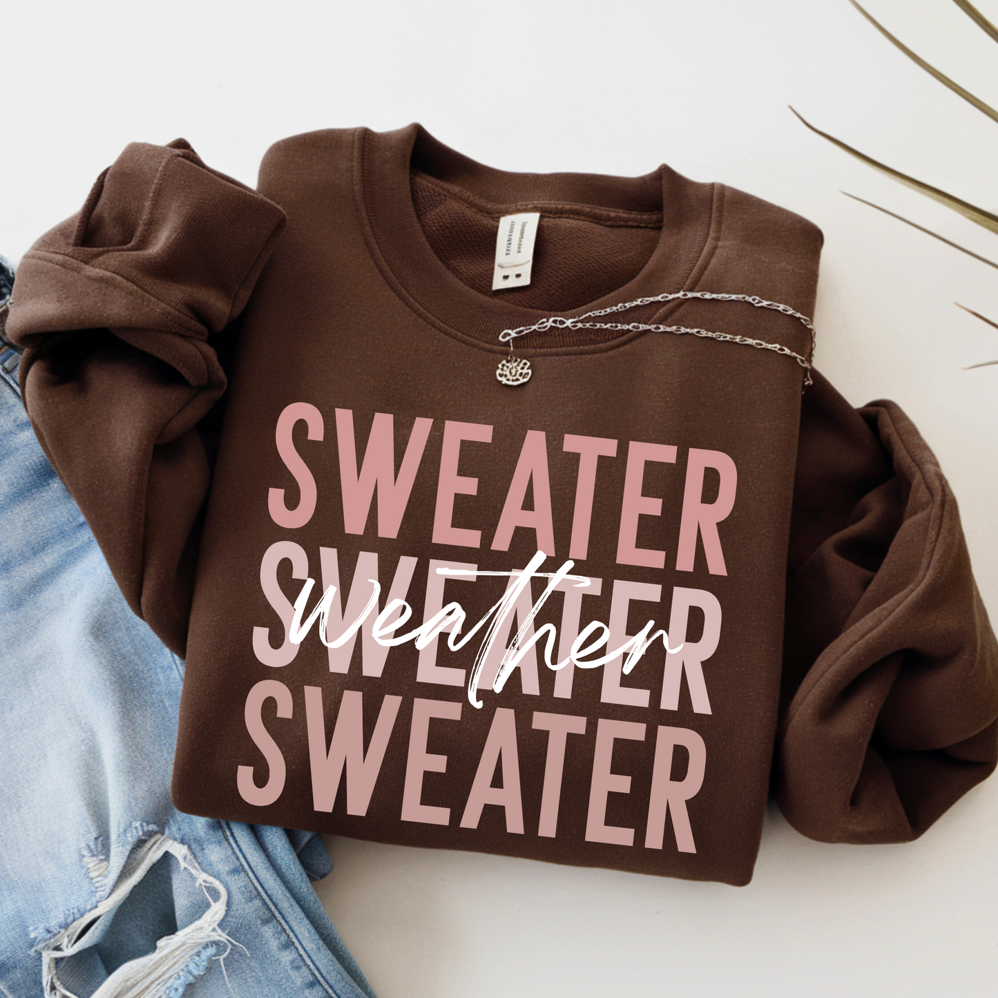 Brown Sweater Weather sweatshirt folded with necklace on top, styled with ripped jeans. Cozy unisex crewneck for fall and winter outfits, perfect for pumpkin patch trips, holiday photos, or lounging.