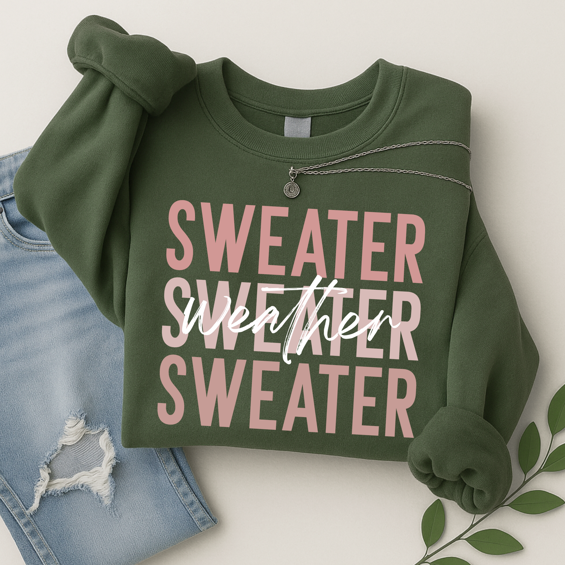 Military green Sweater Weather sweatshirt folded with necklace, styled with ripped jeans. Cozy unisex crewneck, perfect for women’s fall outfits, pumpkin patch visits, and autumn layering.