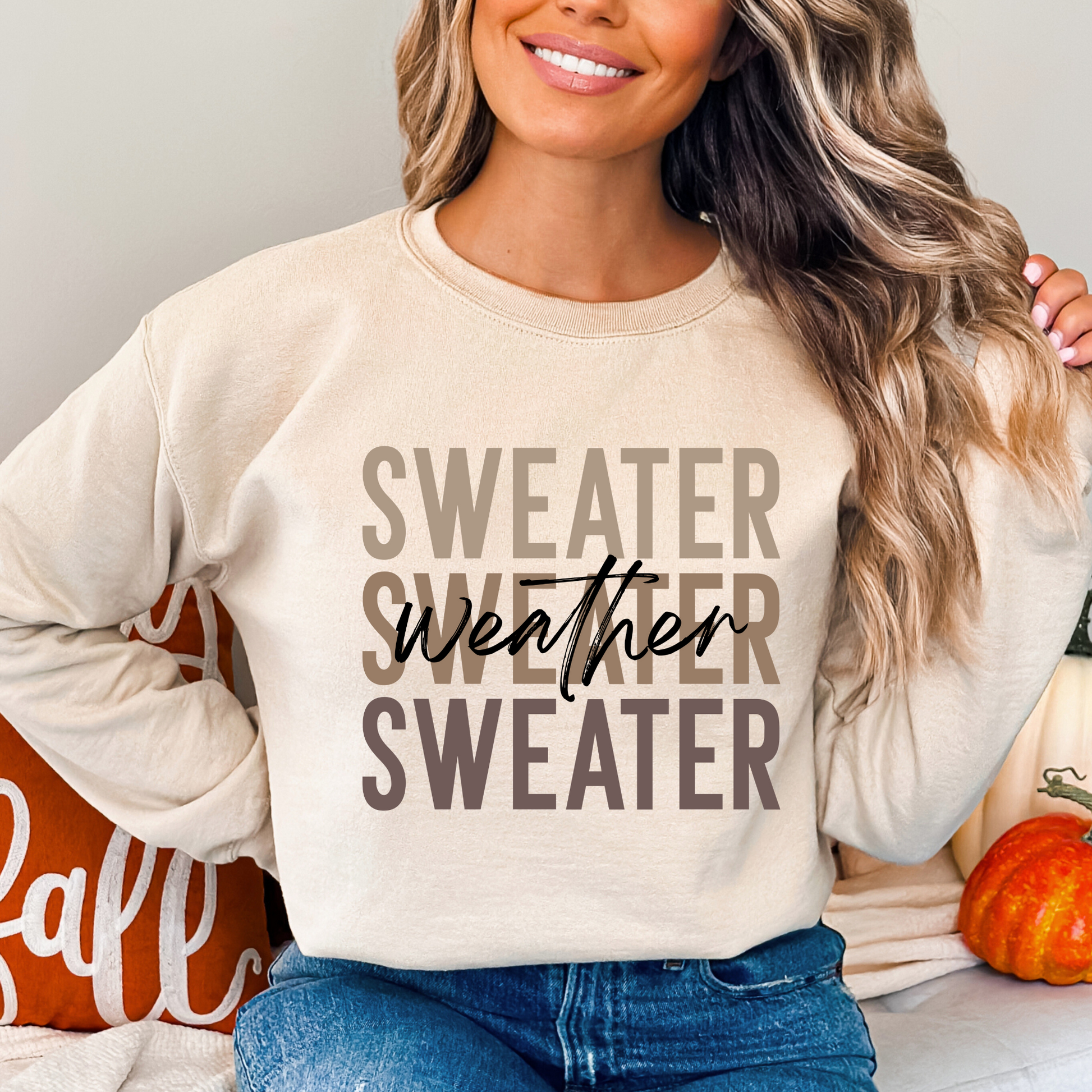 Woman sitting with pumpkins wearing a sand-colored Sweater Weather sweatshirt. Cozy fall fashion pullover ideal for autumn casual outfits and holiday looks.
