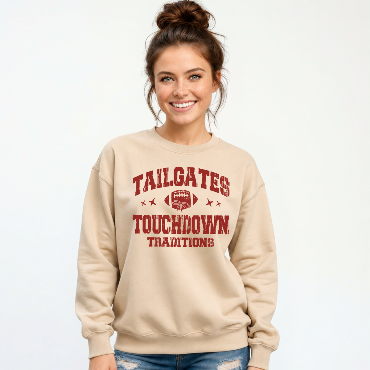 Sand crewneck sweatshirt featuring the Tailgates & Touchdown Traditions graphic, worn by a smiling woman in jeans.