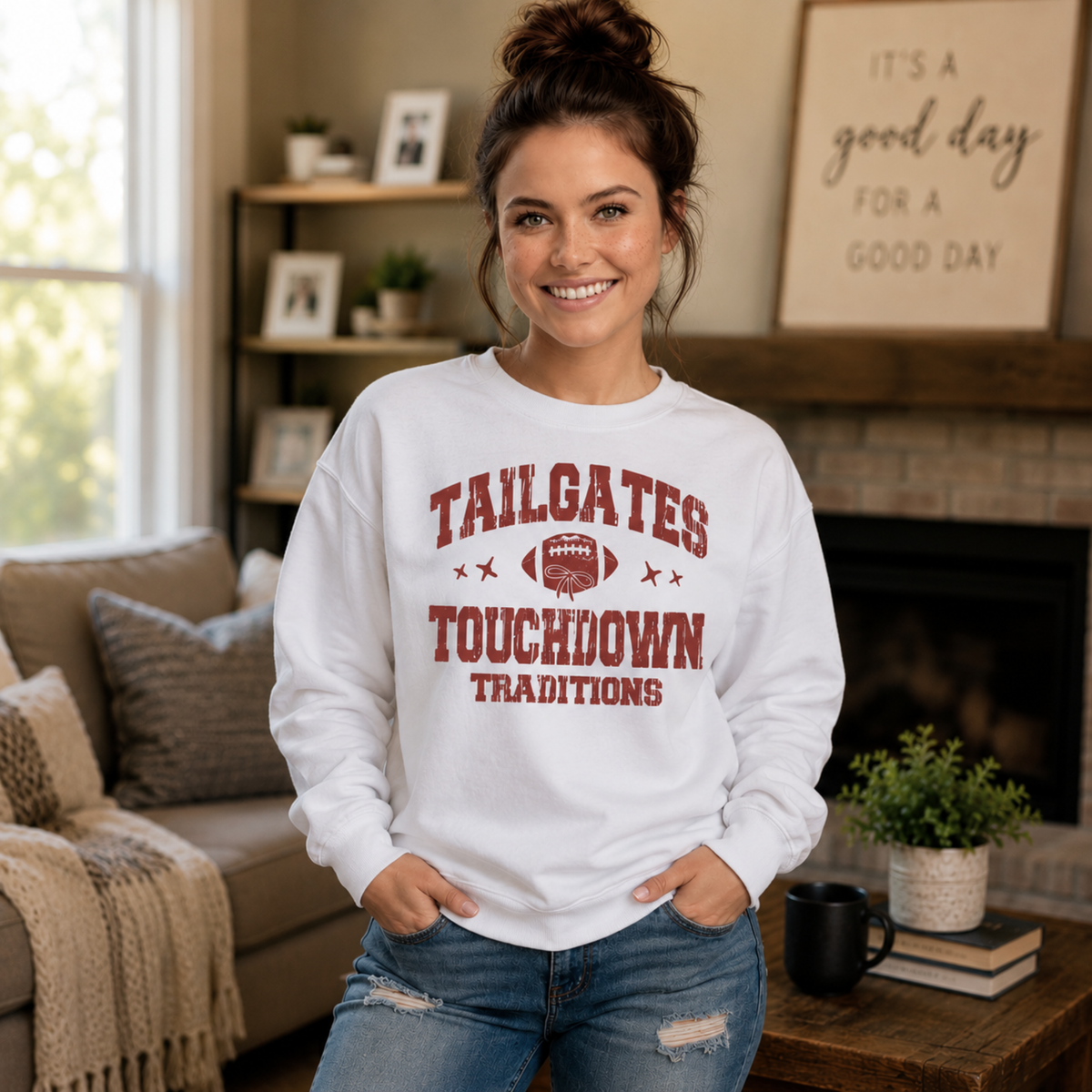 Woman wears a white Tailgates & Touchdown Traditions sweatshirt in a cozy living room lifestyle scene.