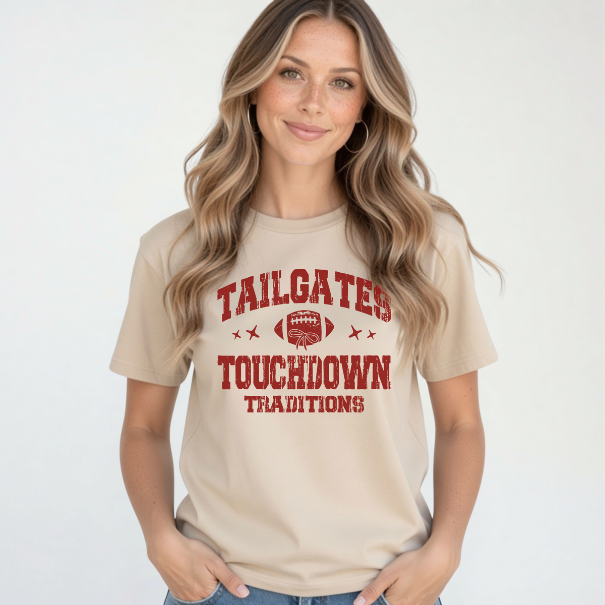 Sand T-shirt featuring the red Tailgates & Touchdown Traditions football graphic, styled on a woman with blue jeans.