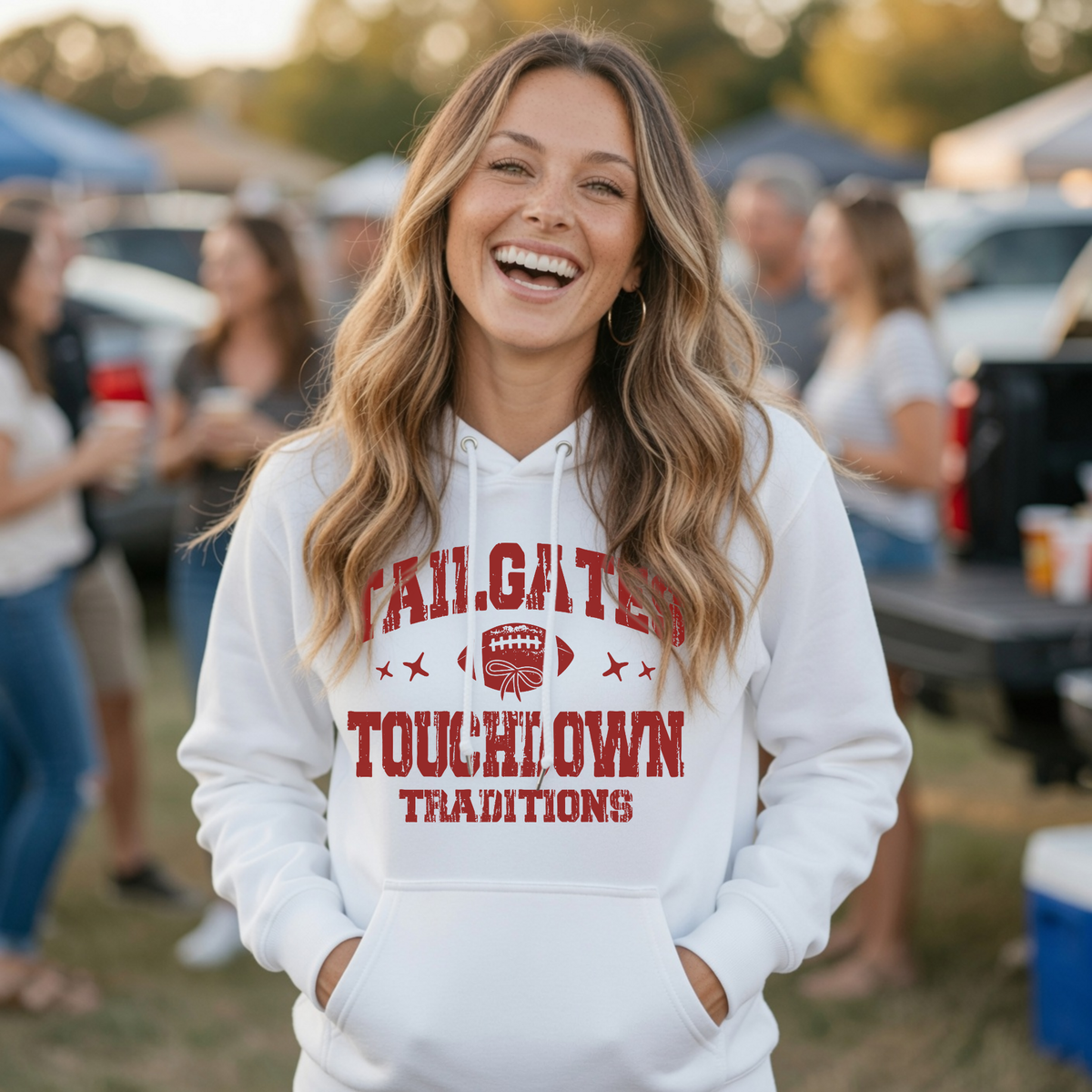 Woman in a white Tailgates & Touchdown Traditions hoodie smiles at an outdoor football tailgate gathering.