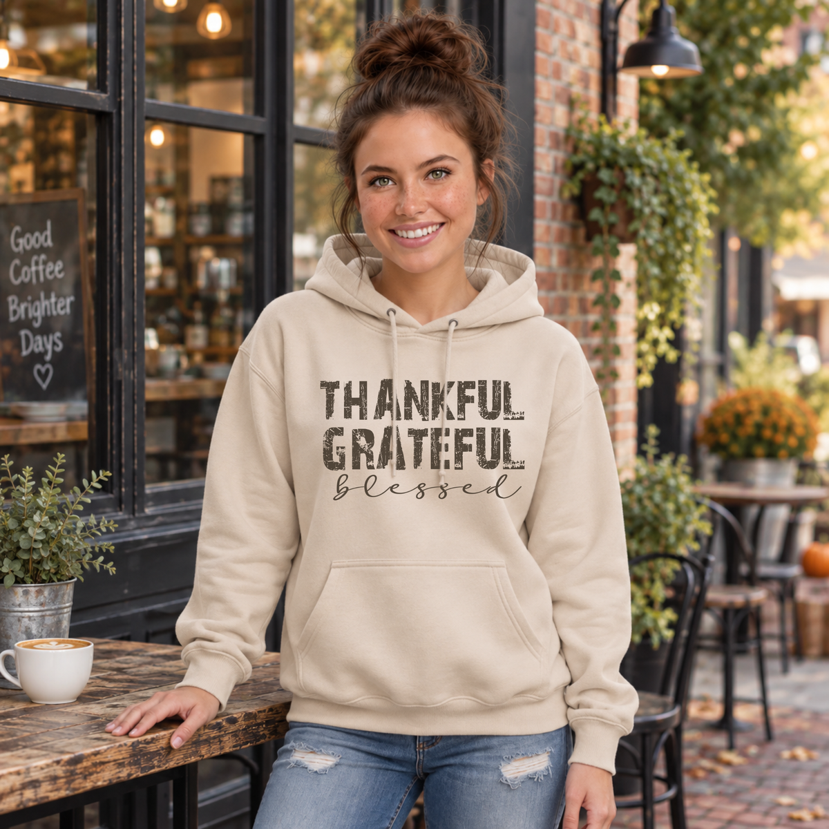 Woman wearing a sand hoodie with Thankful Grateful Blessed lettering while leaning on an outdoor cafe counter.