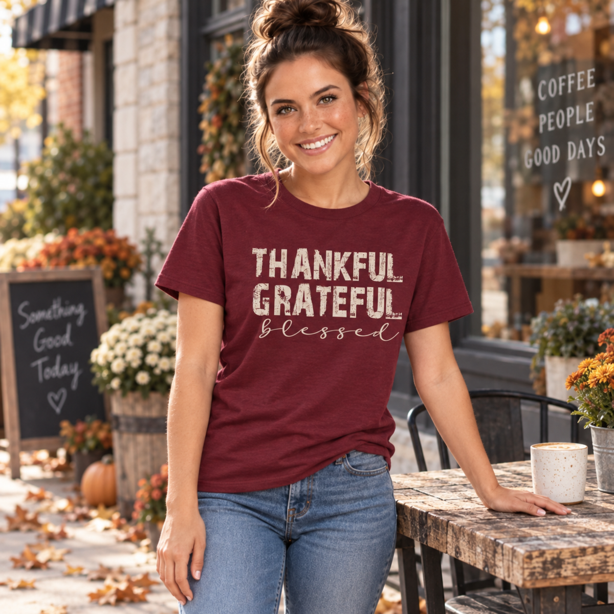 Woman wearing a maroon T-shirt with Thankful Grateful Blessed text beside a cafe table decorated for fall.