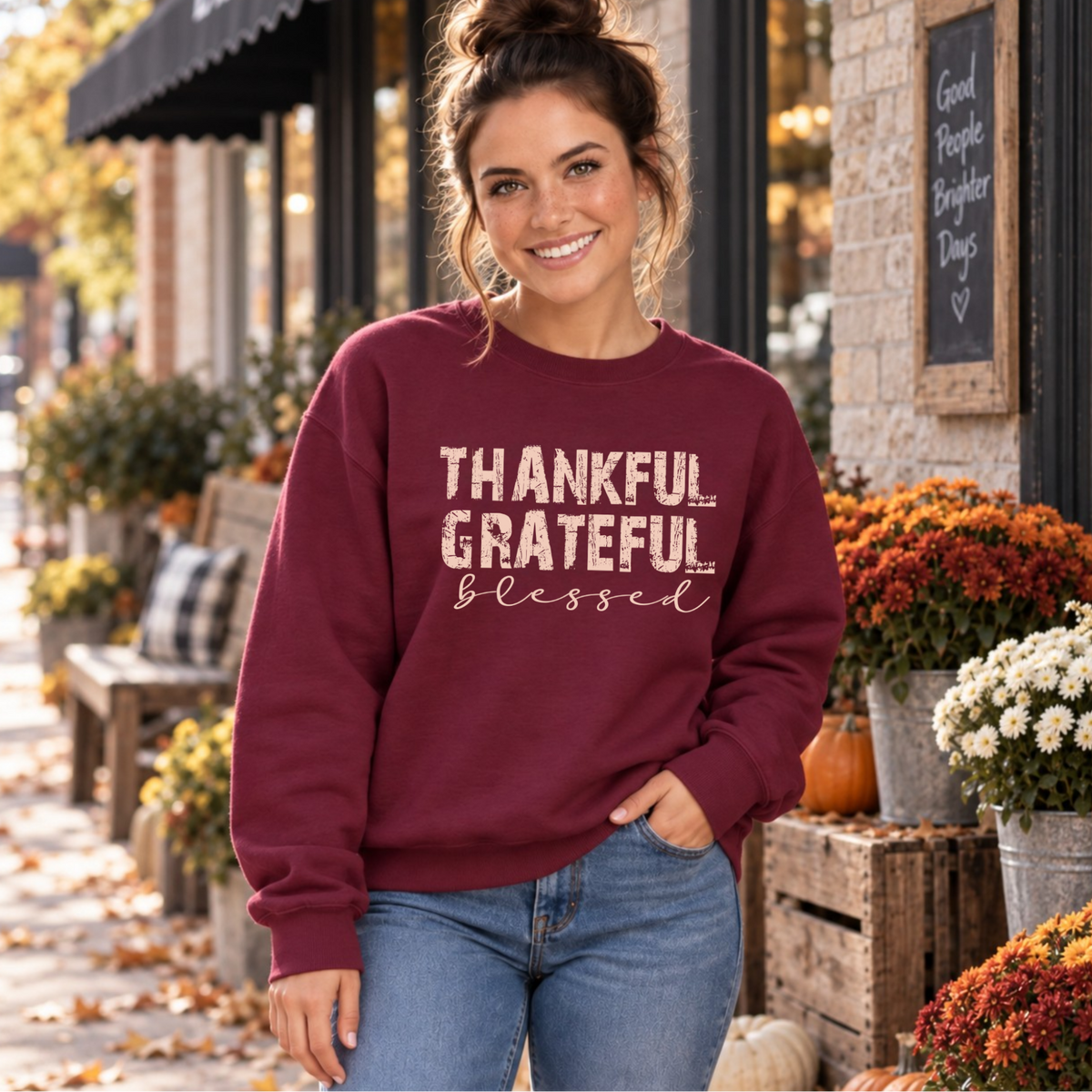 Woman wearing a maroon crewneck sweatshirt with Thankful Grateful Blessed lettering beside fall flowers and pumpkins.