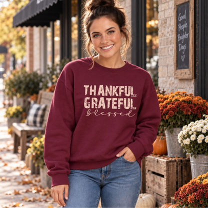 Woman wearing a maroon crewneck sweatshirt with Thankful Grateful Blessed lettering beside fall flowers and pumpkins.