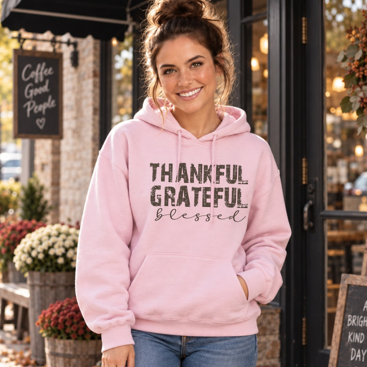 Woman wearing a light pink hoodie with distressed Thankful Grateful Blessed text outside a fall coffee shop.