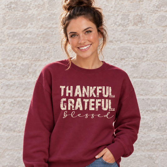 Woman wearing a maroon sweatshirt with Thankful Grateful Blessed lettering against a plain light stone wall.