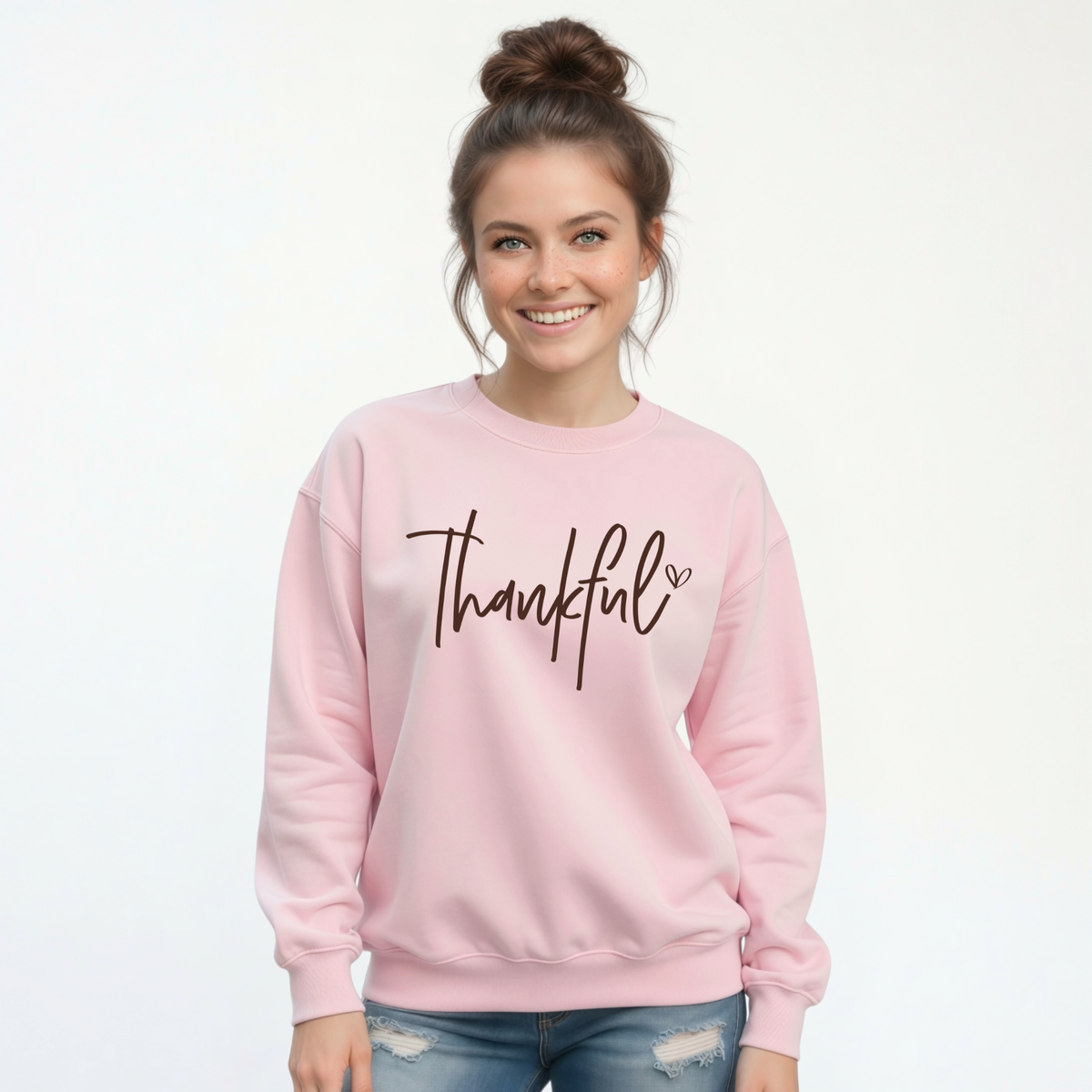 Woman modeling a light pink crewneck sweatshirt with a brown Thankful script design and distressed jeans