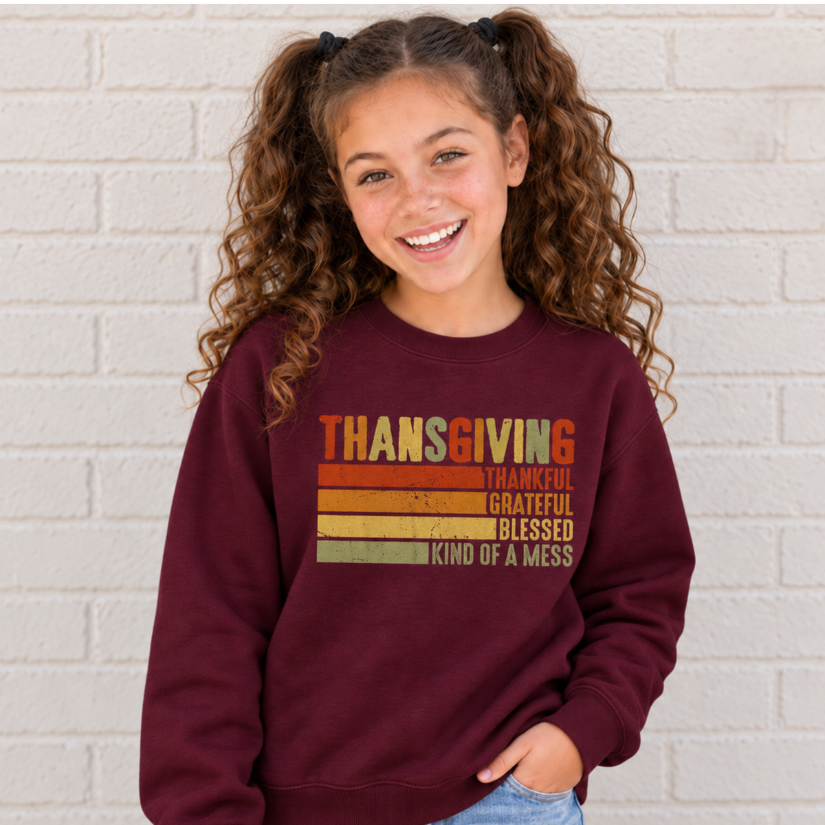 Girl wearing a maroon youth sweatshirt with retro Thanksgiving Kind of a Mess design against a white brick wall.
