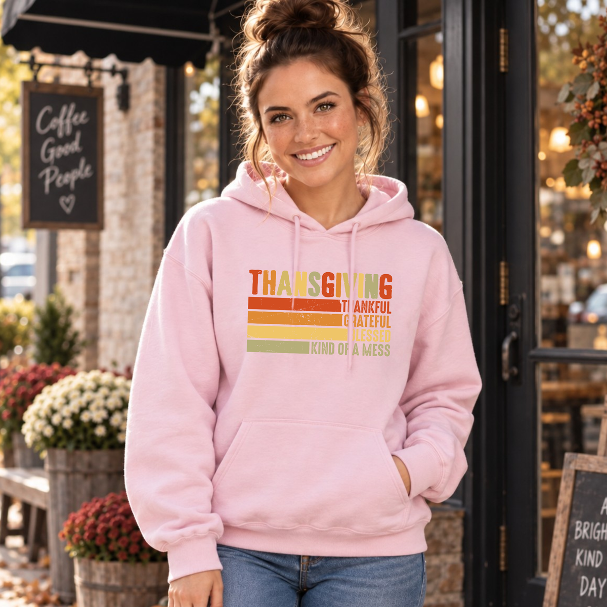Woman wearing a light pink hoodie with retro Thanksgiving Thankful Grateful Blessed Kind of a Mess design outside a cafe.