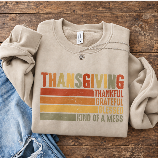 Folded sand sweatshirt with retro Thanksgiving Thankful Grateful Blessed Kind of a Mess design on a rustic wood table.