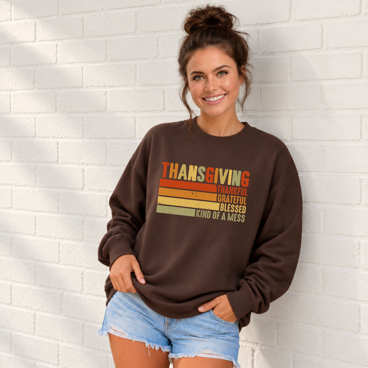 Woman wearing a brown sweatshirt with retro Thanksgiving Thankful Grateful Blessed Kind of a Mess design by a white brick wall.