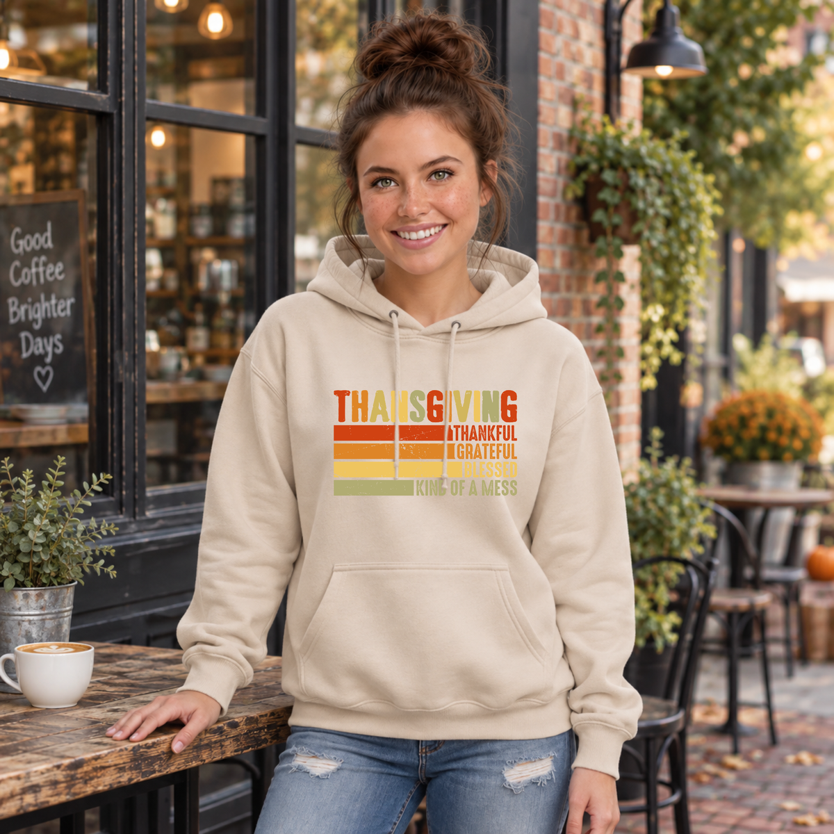Woman wearing a sand hoodie with retro Thanksgiving Thankful Grateful Blessed Kind of a Mess design outside a cafe.
