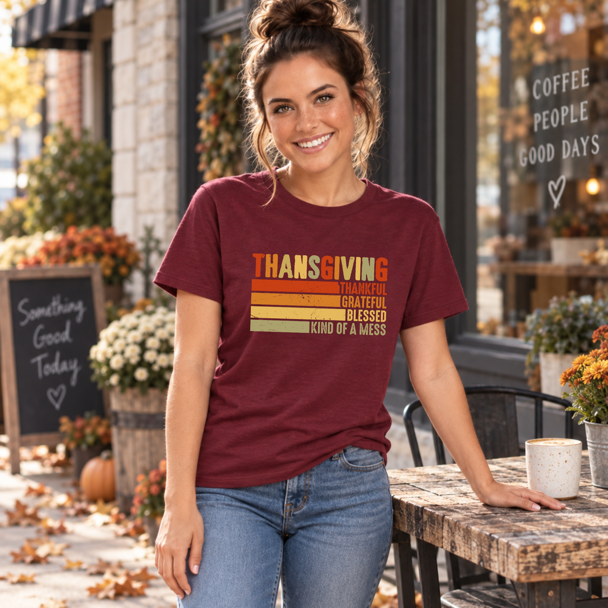 Woman wearing a maroon T-shirt with retro Thanksgiving Thankful Grateful Blessed Kind of a Mess design by a cafe table.