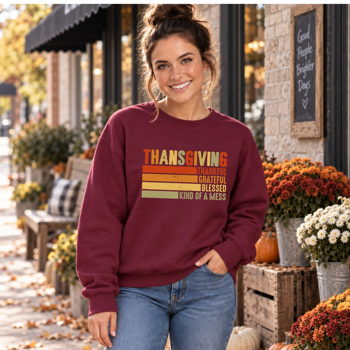 Woman wearing a maroon sweatshirt with retro Thanksgiving Kind of a Mess design beside fall flowers and pumpkins.