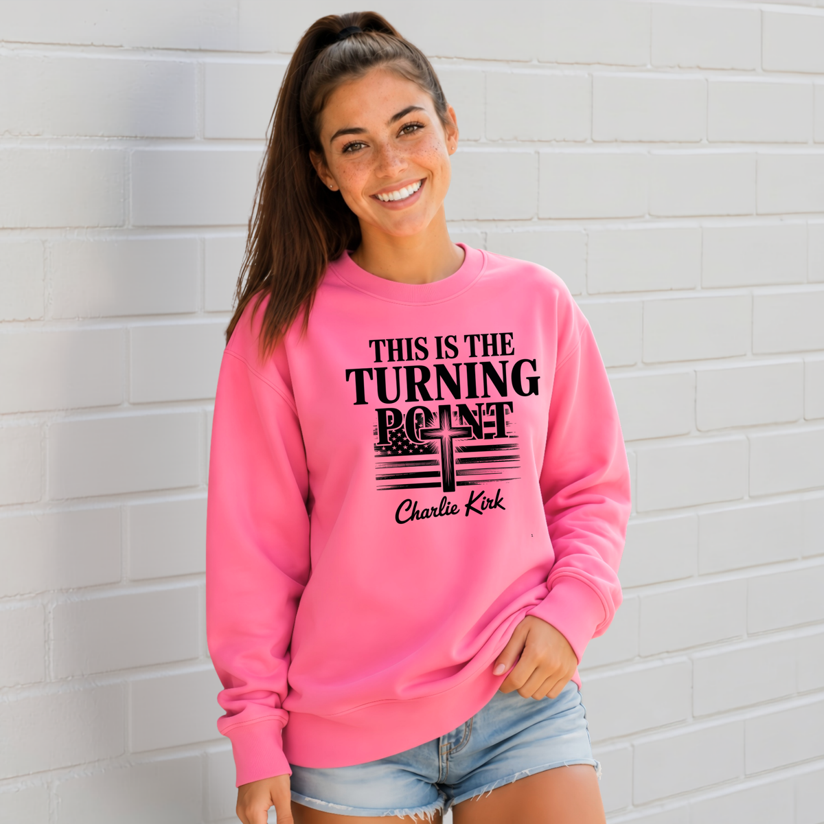 Woman wearing a bright pink crewneck sweatshirt with black This Is the Turning Point cross graphic.