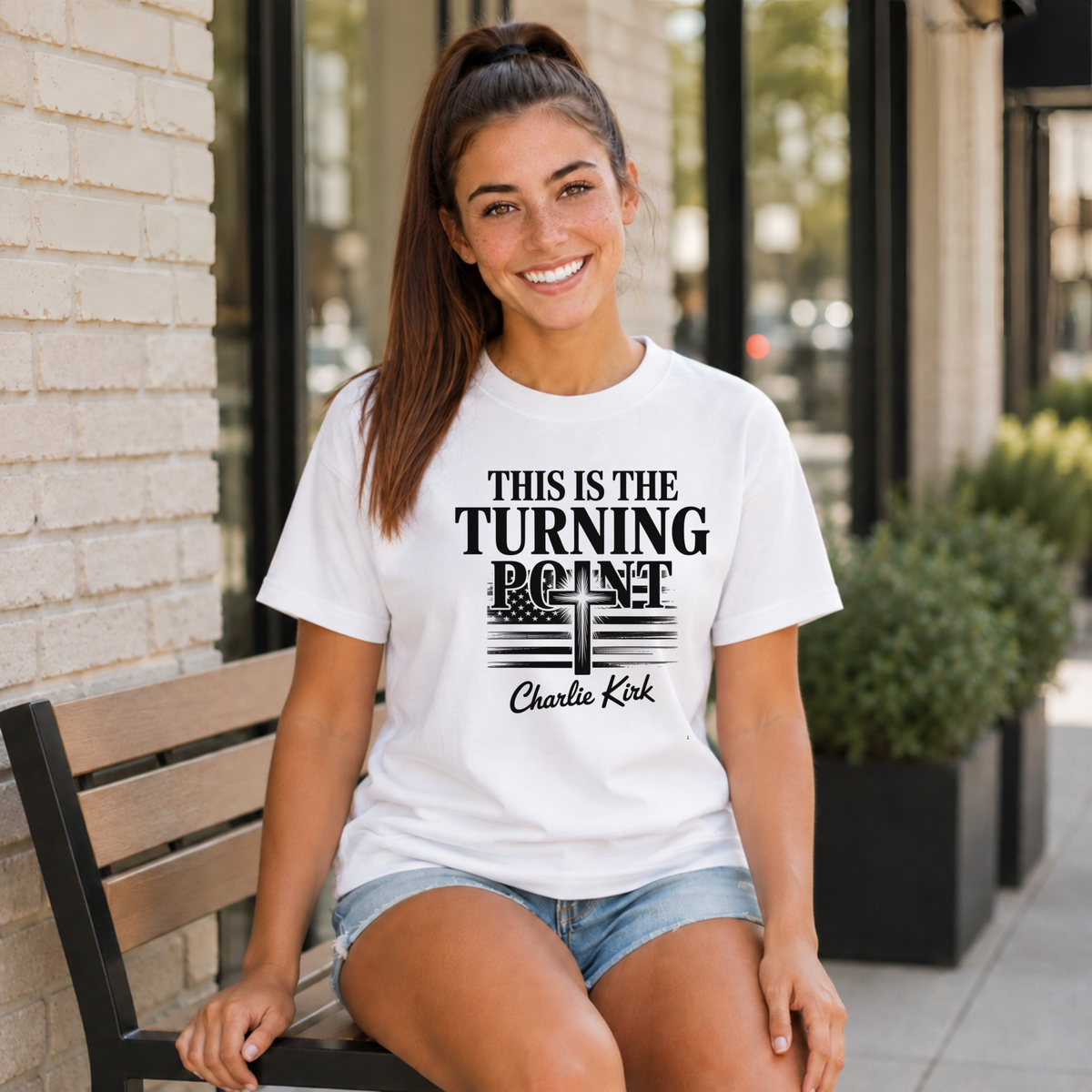 Woman seated outside wearing a white T-shirt with black This Is the Turning Point cross graphic.