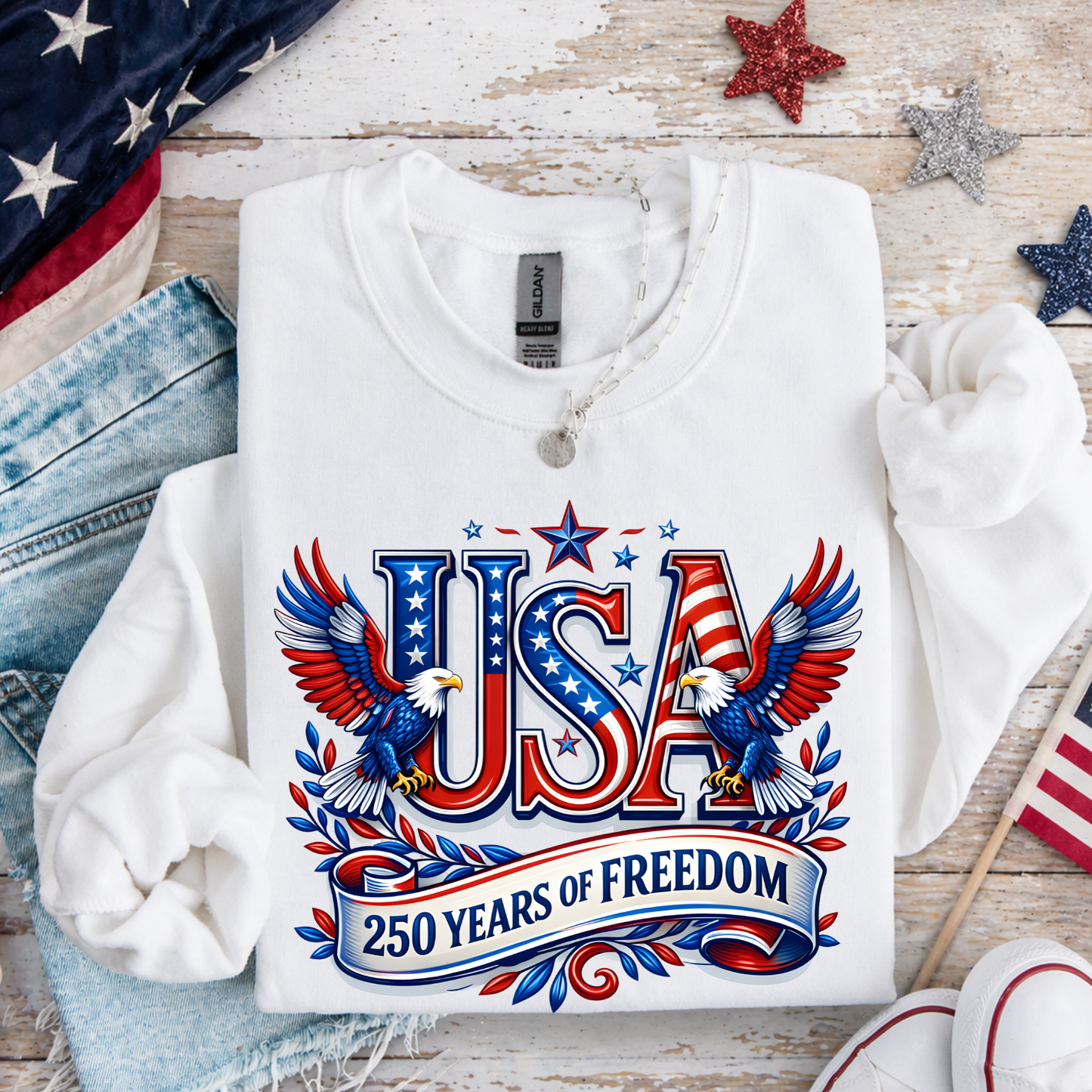 White sweatshirt with 'USA 250 Years of Freedom' design on a wooden surface with American flags and stars.
